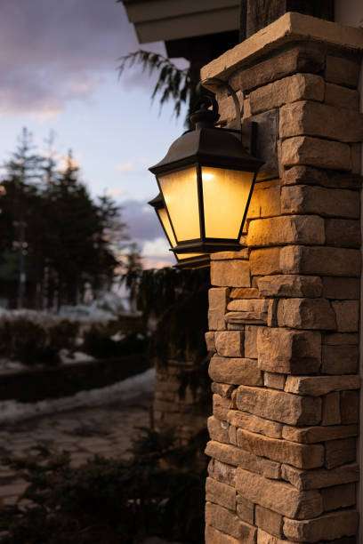 Lantern-style wall light illuminating a rustic stone pillar at dusk, with snowy trees.
