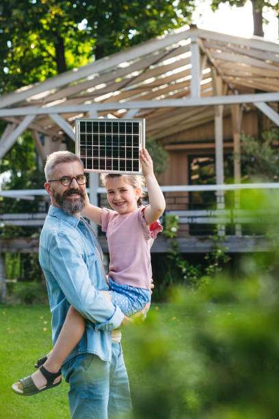 Father and daughter hold outdoor solar light panel near greenhouse