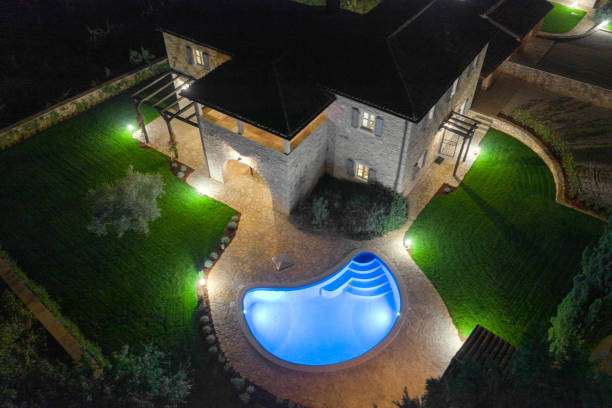 Stunning aerial night view of a mansion, pool, and garden illuminated by elegant wall light fixtures.